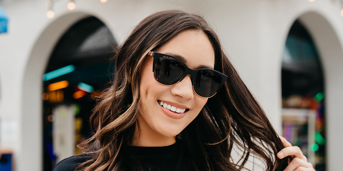 A close up, angled image of a smiling woman in a black shirt wearing the Americano Blenders Sunglasses, showcasing a front view of the crystal gray and solid black tortoise pattern frame and polarized silver mirrored lenses of the sunglasses.