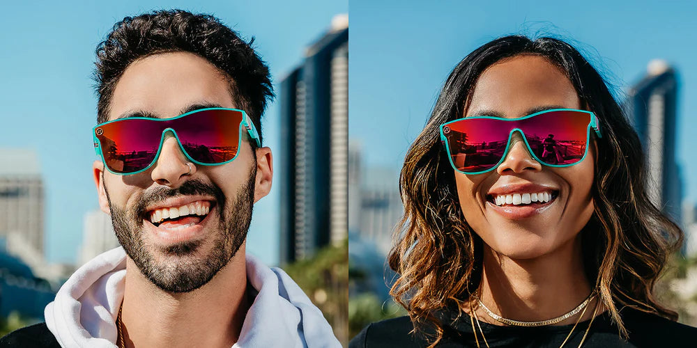A close up image of a man's and woman's smiling faces looking straight wearing the Dance Electric Blenders Sunglasses, showcasing a front view of the gloss crystal teal frame and polarized hot pink mirrored lenses of the sunglasses.