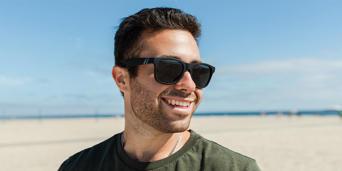 A close up image of a smiling man in a green shirt looking left, wearing the Deep Space X2 Blenders Sunglasses, showcasing a front, angled view of the matte black frame and polarized smoke lenses of the sunglasses.