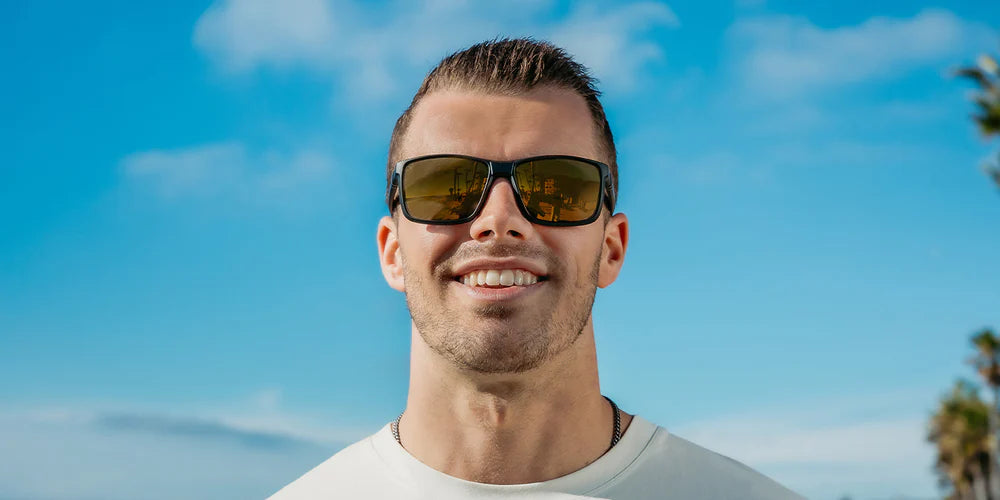 A close up image of a man smiling looking straight wearing the Matte Black Blenders Sunglasses, showcasing a front view of the matte black frame and polarized copper mirrored lenses of the sunglasses.