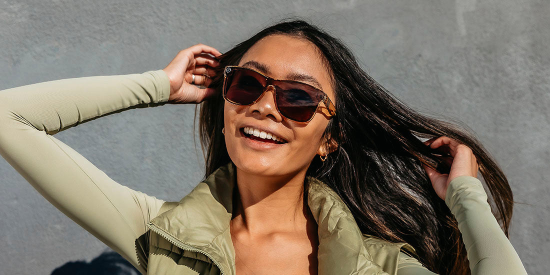 A close up, image of a smiling woman in a light green vest and shirt wearing the Mojave Mirage Blenders Sunglasses, showcasing a front view of the gloss crystal tan frames and polarized amber lenses of the sunglasses.