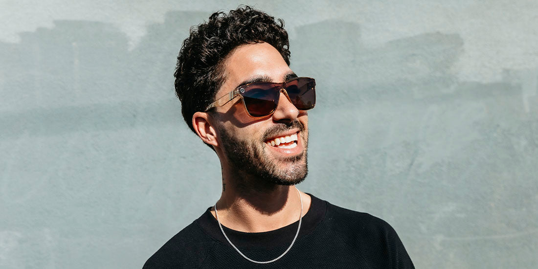 A close up, angled image of a smiling man in a black shirt wearing the Mojave Mirage Blenders Sunglasses, showcasing a front, side view of the gloss crystal tan frames and polarized amber lenses of the sunglasses.