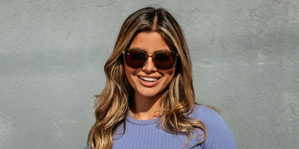 A close up image of a smiling woman in a blue shirt wearing the Mojave Moon Blenders Sunglasses, showcasing a front view of the crystal tan frame and PureBlend™ amber lenses of the sunglasses.