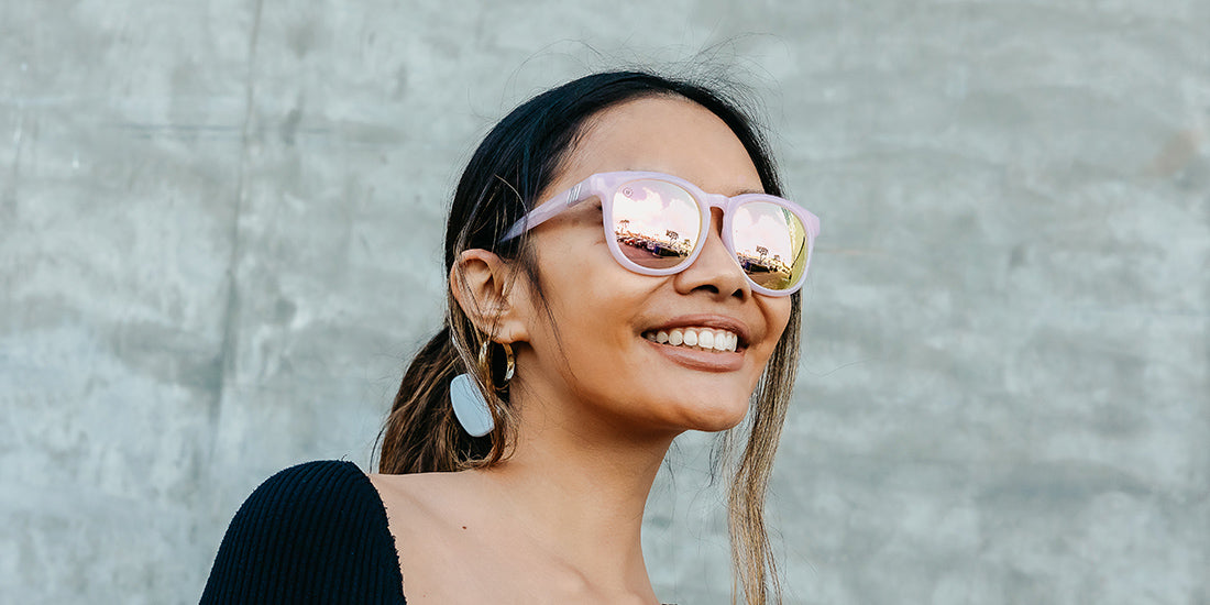 A close up image of a smiling woman in a black shirt wearing the Pink Rose Blenders Sunglasses, showcasing a front, angled view of the glossed milky pink frame and polarized PureBlend™ pink mirrored lenses of the sunglasses.