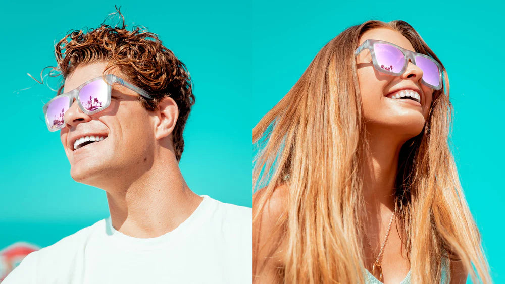 A close up image of a man's and woman's smiling faces looking in the opposite directions wearing the Pretty Gangsta' Blenders Sunglasses, showcasing a front view of the gloss crystal clear gray and pink tortoise frame and polarized pink mirrored lenses of the sunglasses.