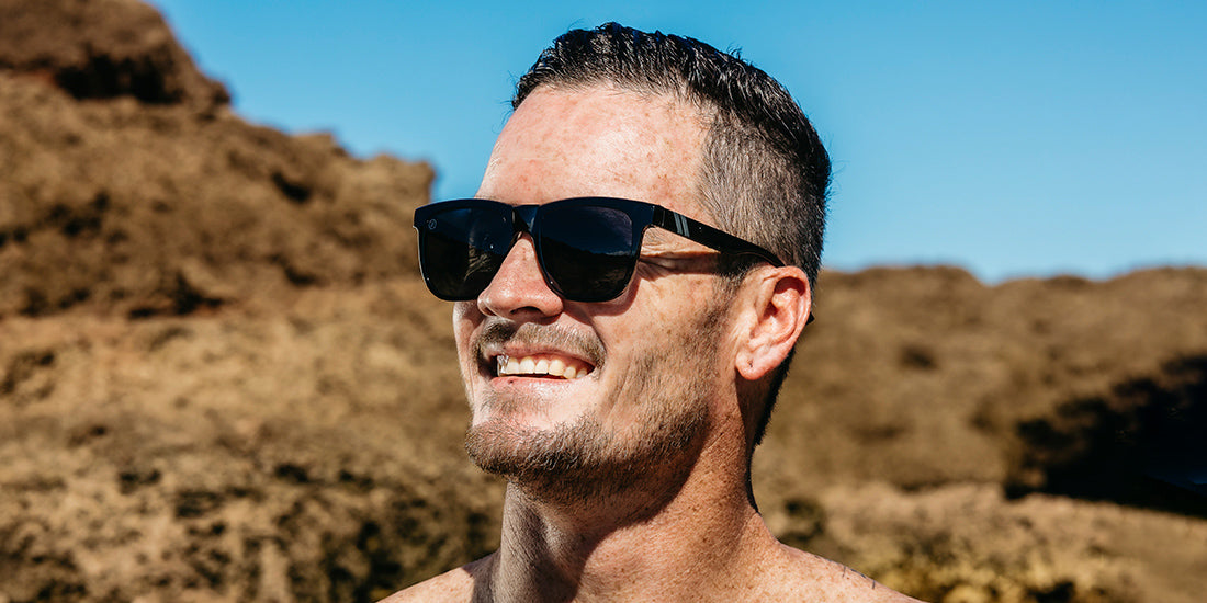 A close up image of a smiling man wearing the Shadow Cruise Blenders Sunglasses, looking right showcasing an angled front view of glossy black frame and PureBlend™ smoke black lenses of the sunglasses.