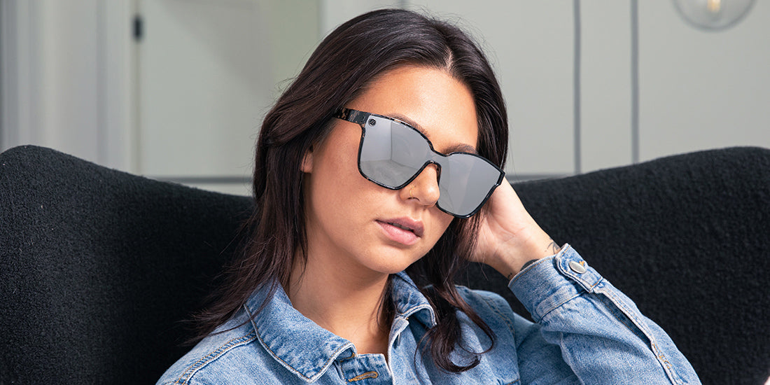 A close up, image of a woman in a denim jacket wearing the Sterling Lady Blenders Sunglasses, showcasing a front view of the crystal gray and solid black tortoise pattern frames and polarized silver mirrored lenses of the sunglasses. 