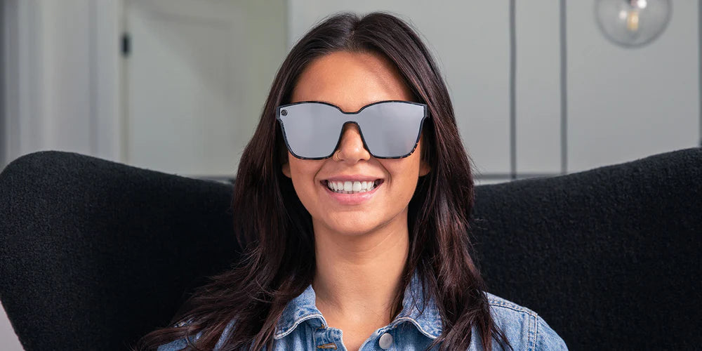 AA close up, image of a smiling woman in a denim jacket wearing the Sterling Lady Blenders Sunglasses, showcasing a front view of the crystal gray and solid black tortoise pattern frames and polarized silver mirrored lenses of the sunglasses. 
